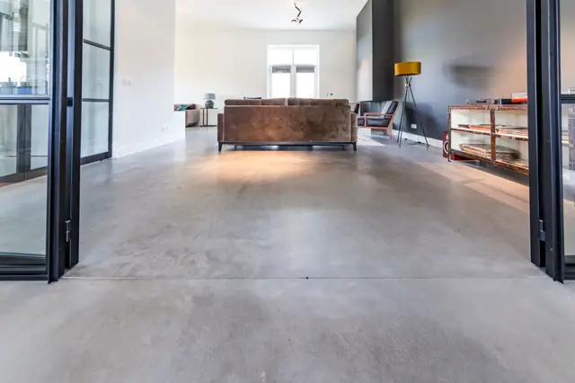 Ruime, lichte woonkamer met een naadloze vloer in warm grijs. Groot raam met zwarte frames, bruine designmeubels en minimalistisch ingerichte ruimte met perfecte afwerking.
