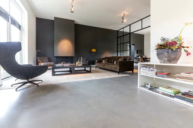 Ruime, open woonkamer met strakke architectuur, grote ramen en natuurlijk licht. De naadloze vloer loopt naadloos door, perfecte afwerking met donkere accentwanden en design meubilair.