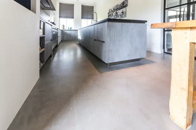 Ruime, lichte keuken met strak design. Naadloze vloer loopt elegant door, aangevuld met grijze keukeneiland en witte wanden. Natuurlijk licht van groot raam, minimalistisch interieur.