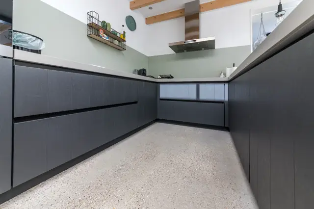 Strakke grijze keukenkastjes met zwarte greeplijnen tegen een lichte wand met zachtgroen accent. De naadloze vloer in licht terrazzo-achtig materiaal loopt vogelsvlucht door de ruimte, wat een schoon en minimalistisch eindresultaat toont.