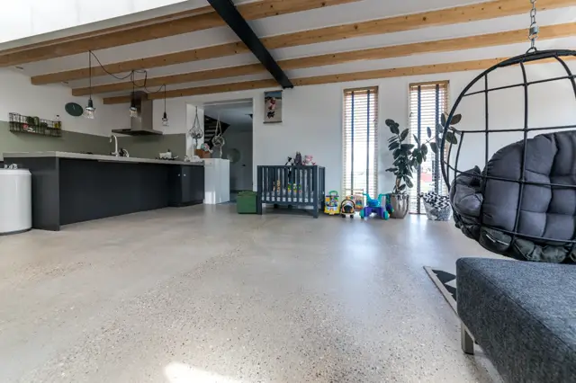 Grootschalige open woon- en keukenruimte met een lichte, naadloze vloer. De ruimte combineert moderne keukenkasten met houten dakbalken en veel natuurlijk licht via grote ramen.
