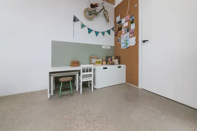 Een rustig ingerichte kinderkamer met een grijsbeige naadloze vloer als basis. De ruimte toont een gestructureerde opstelling met wit bureau, groen accent aan de wand en opbergruimte, gecombineerd met persoonlijke decoratie.