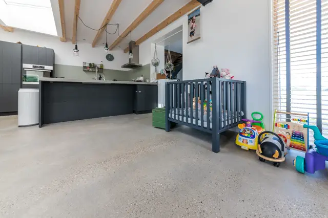 Ruime, lichte woon- en keukenruimte met houten balkenplafond, zwarte keukenkasten en een naadloze vloer. De ruimte wordt echter significant beperkt door speelgoed en kindermeubilair.
