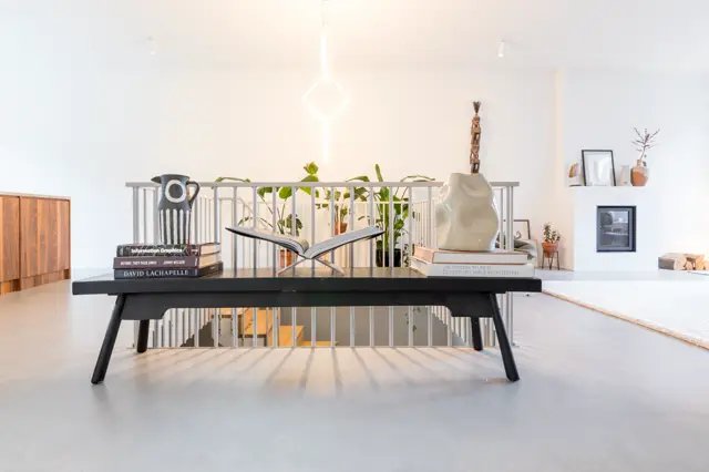 Lichte, open ruimte met een strakke naadloze vloer in warm grijs. Een design salontafel met decoratieve objecten staat centraal; natuurlijk licht valt in rechte lijnen door grote ramen. Witte muren, zwevende planken en sfeervolle styling met planten en boeken.