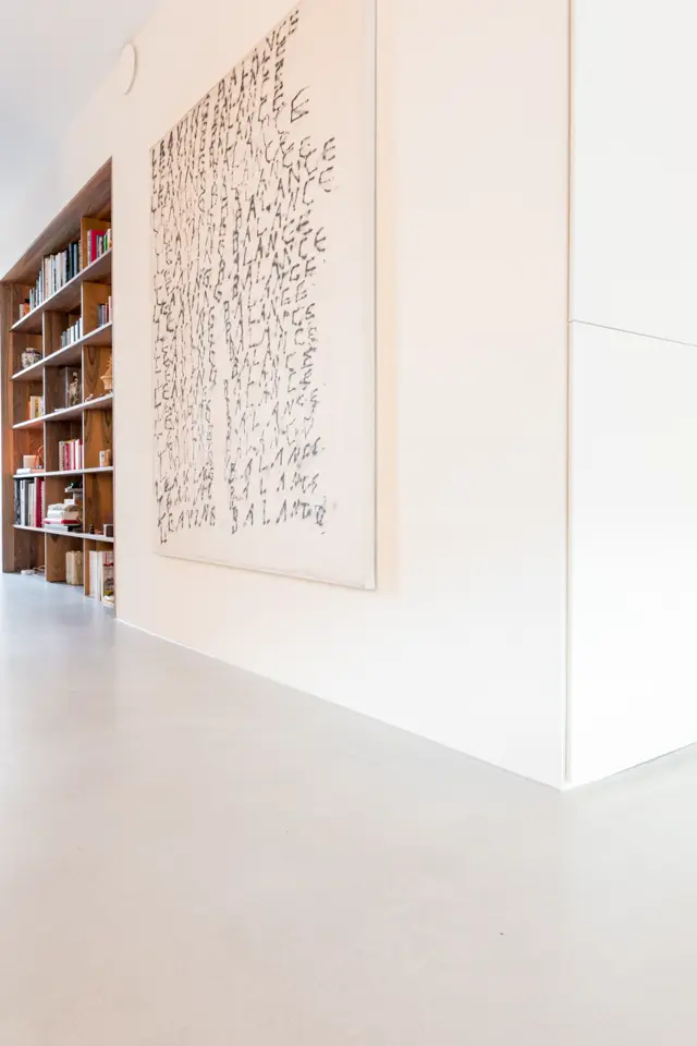 Schone, lichte hal met een strakke crèmekleurige naadloze wand, voorzien van abstract tekstwerk. Houten boekenkasten met witte schappen creëren warmte en contrast.