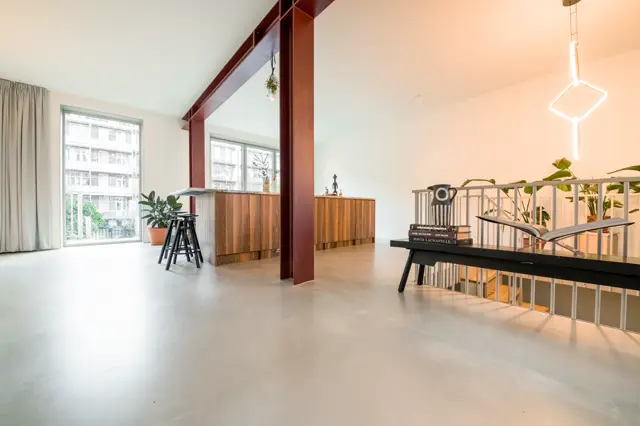 Ruime, lichte woonruimte met een strakke naadloze vloer in licht grijs. Het interieur combineert industriële elementen met moderne design, grote ramen zorgen voor natuurlijk licht.