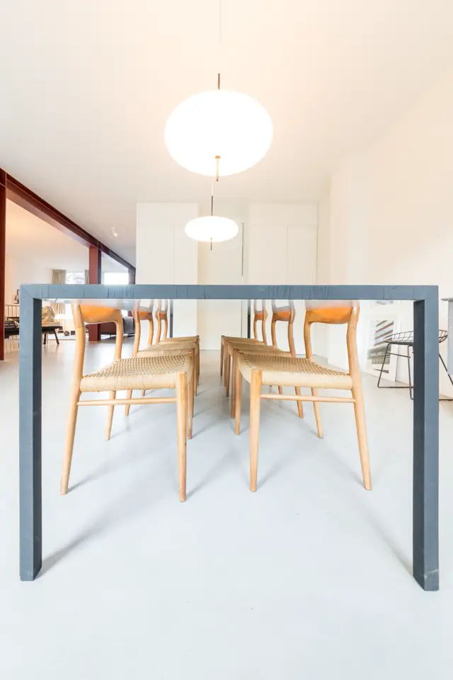 Strakke, lichte eetkamer met een naadloze vloer in warm wit. Houten tafel met natuurlijke stoelen, gekenmerkt door schone lijnen en perfect afgewerkte wanden. Moderne hangende verlichtingselementen zorgen voor diffuus licht.