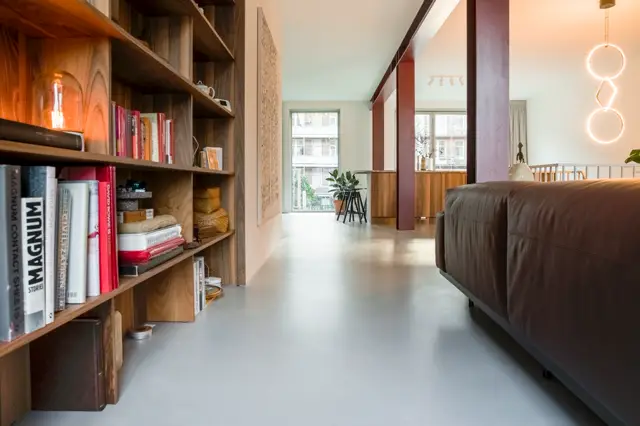 Ruime, lichte woonruimte met een strakke grijze naadloze vloer, houten inbouwkasten en minimalistische styling. Grote ramen zorgen voor natuurlijk licht en rust.