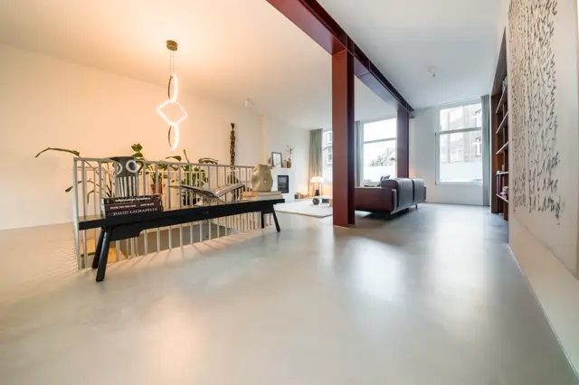 Ruime, lichte woonruimte met industriële accenten en een naadloze grijze vloer. De open lay-out wordt gedefinieerd door het contrast van warm licht en strakke lijnen.