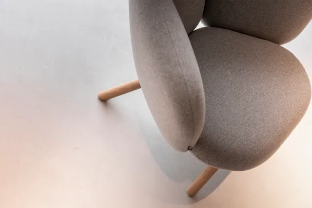 Een elegante, ronde stoel met grijsbeige bekleding en houten poten staat op een schone, lichte naadloze vloer. Het beeld straalt minimalistische rust en verfijnde proporties uit.