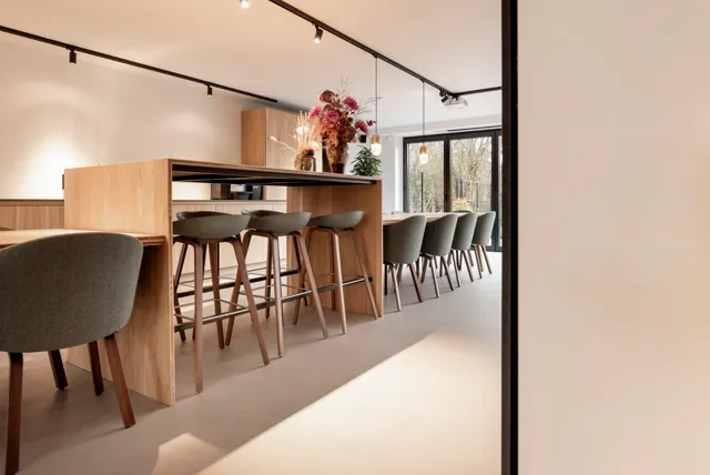 Minimalistisch ingerichte ruimte met houten keukeneiland, barkrukken en eettafel. Grote ramen brengen natuurlijk licht naar binnen. De naadloze vloer loopt ononderbroken door de hele ruimte, ondersteunend voor de strakke, open architectuur.