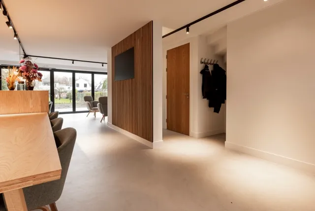 Ruime, licht ingevallen hal met subtiele warme tinten, naadloze vloer en schone geometrische lijnen. Houten accenten en strakke wanden creëren een geruststellende, geordende sfeer.
