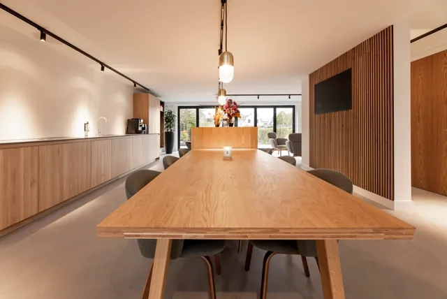 Lichte, strakke openplan ruimte met houten accenten, grote ramen en een naadloze vloer in warme, neutrale tint. De ruimte ademt rust en precisie uit met scherpe lijnen en minimalistisch design.
