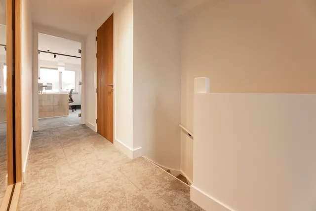 Een strakke, lichte hal met naadloze vloer in warm beige, witte wanden en houten deuren. De ruimte ademt rust en helderheid uit met scherpe lijnen en perfect afgewerkte details.