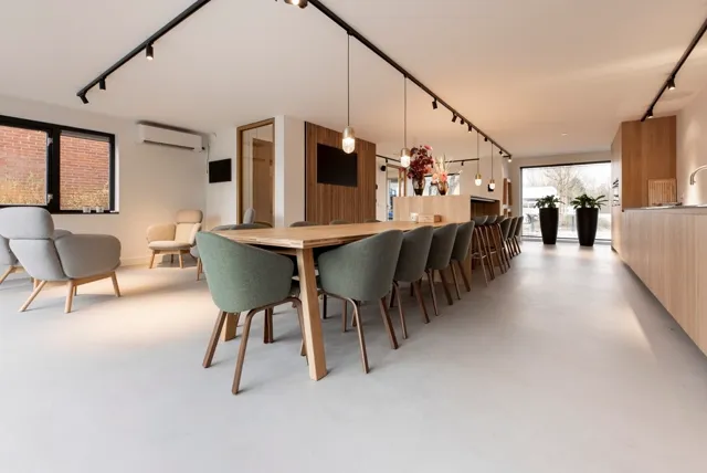 Moderne open werkplek met lange houten tafel, designstoelen en naadloze lichte vloer. Ruim, helderder ingericht met houten accenten en minimalistische inrichting.