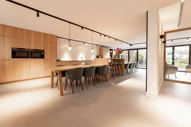 Minimalistisch ontworpen keuken-eetkamer met houtaccenten en een egale, naadloze vloer die de ruimte visueel vergroten. Natuurlijk licht domineert de strakke, opgeruimde setting.