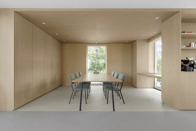 Een strak, licht eetgedeelte met houten wanden en plafond, naadloze lichte vloer, en grote ramen met natuurlijk licht. De ruimte ademt rust en soberheid uit met een eenvoudig tafeltje en vier stoelen.